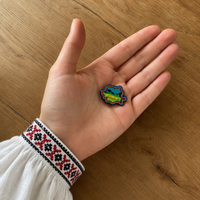 Broșă România CheiFolk ținută în palmă, cu ie tradițională românească și fundal din lemn